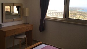 Ein Schlafzimmer mit einem Holztisch, einem Stuhl, einem Spiegel, einer Lampe und einem Fenster mit Blick auf eine Stadtlandschaft.
