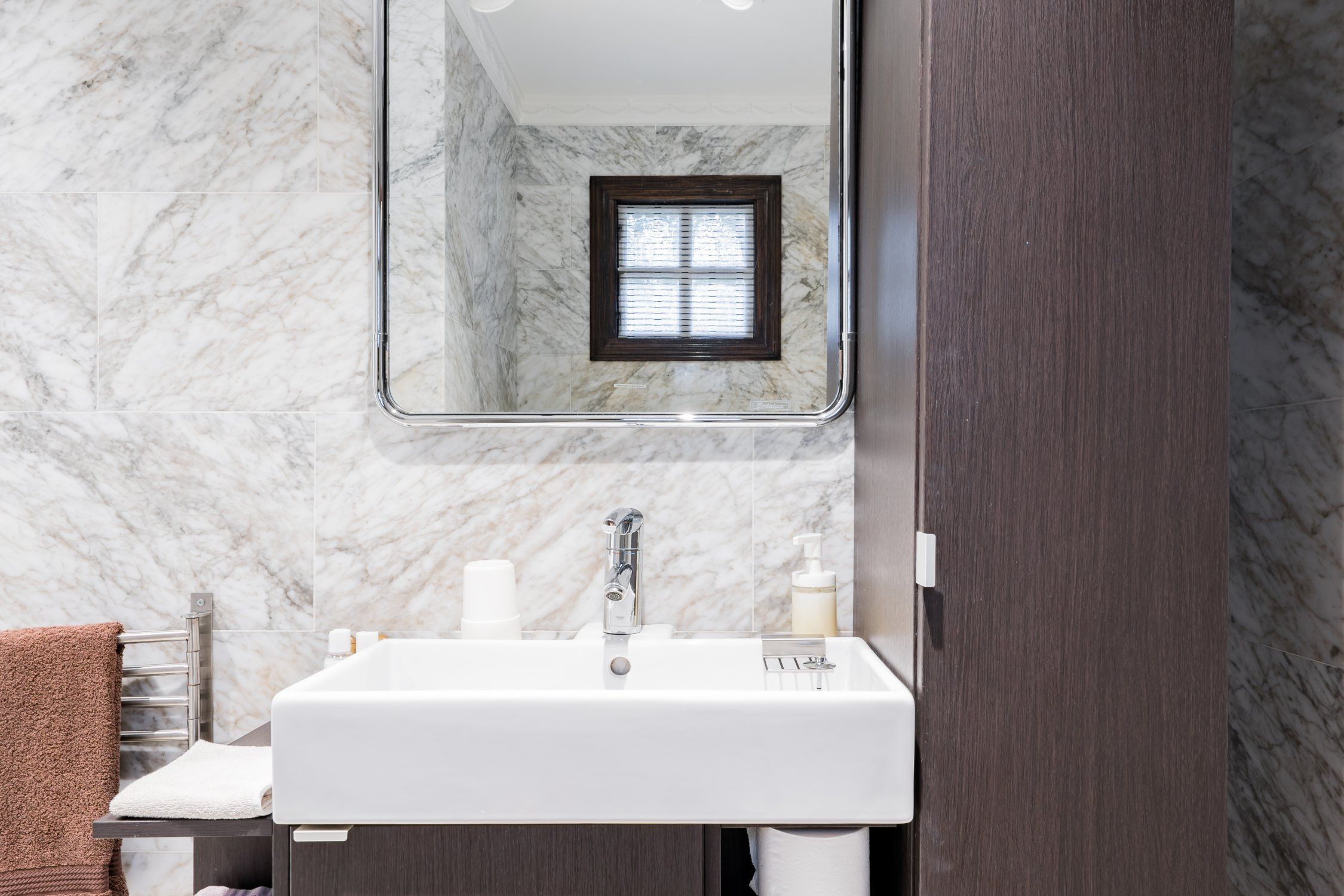 A bathroom with a marble wall, a white sink, a mirror, and a window.