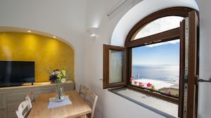 A dining area with a wooden table, chairs, a vase of flowers, a flat-screen TV, and a window with a view of the sea. - Golden Vaults, at few stairs from the road. It is easy to park in the area (Praiano)