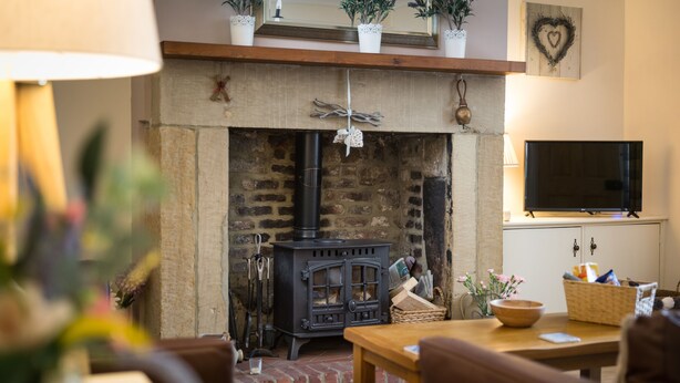 A cozy living room with a stone fireplace, a television mounted on the wall, and a wooden table with a basket and a bowl. - Charming Original Stone Cottage Close To Beaches and Countryside (Alnwick)