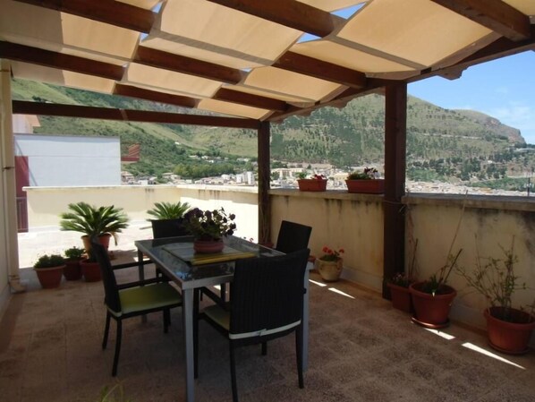 A covered outdoor seating area with a table and chairs, potted plants, and a view of a town and hills in the background.