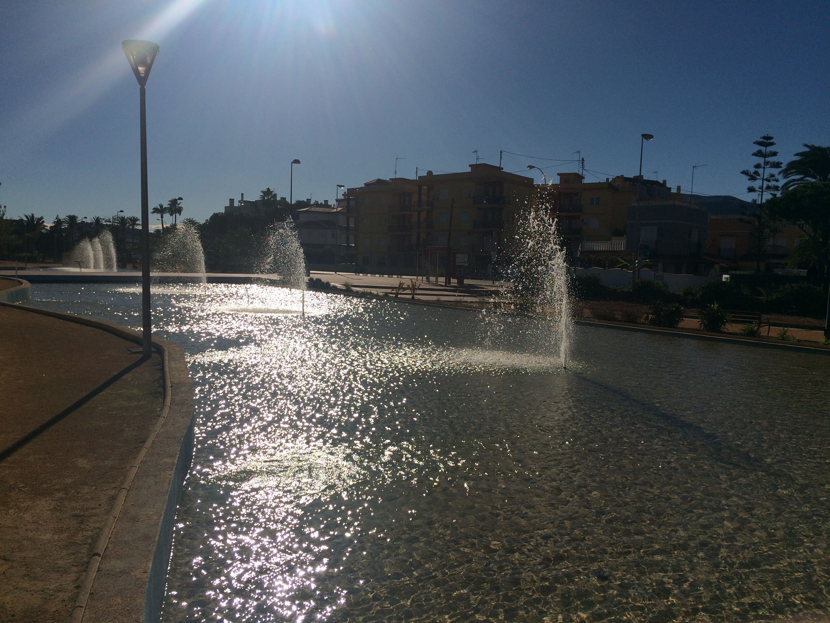 Uma fonte com jatos de água numa praça pública, rodeada por edifícios e um céu claro.
