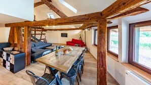Ein geräumiges Zimmer mit einem Holzesstisch, einer Couch und einem roten Sessel. Eine Treppe mit Holzbalken und ein Fenster mit Blick auf Grün sind ebenfalls vorhanden.