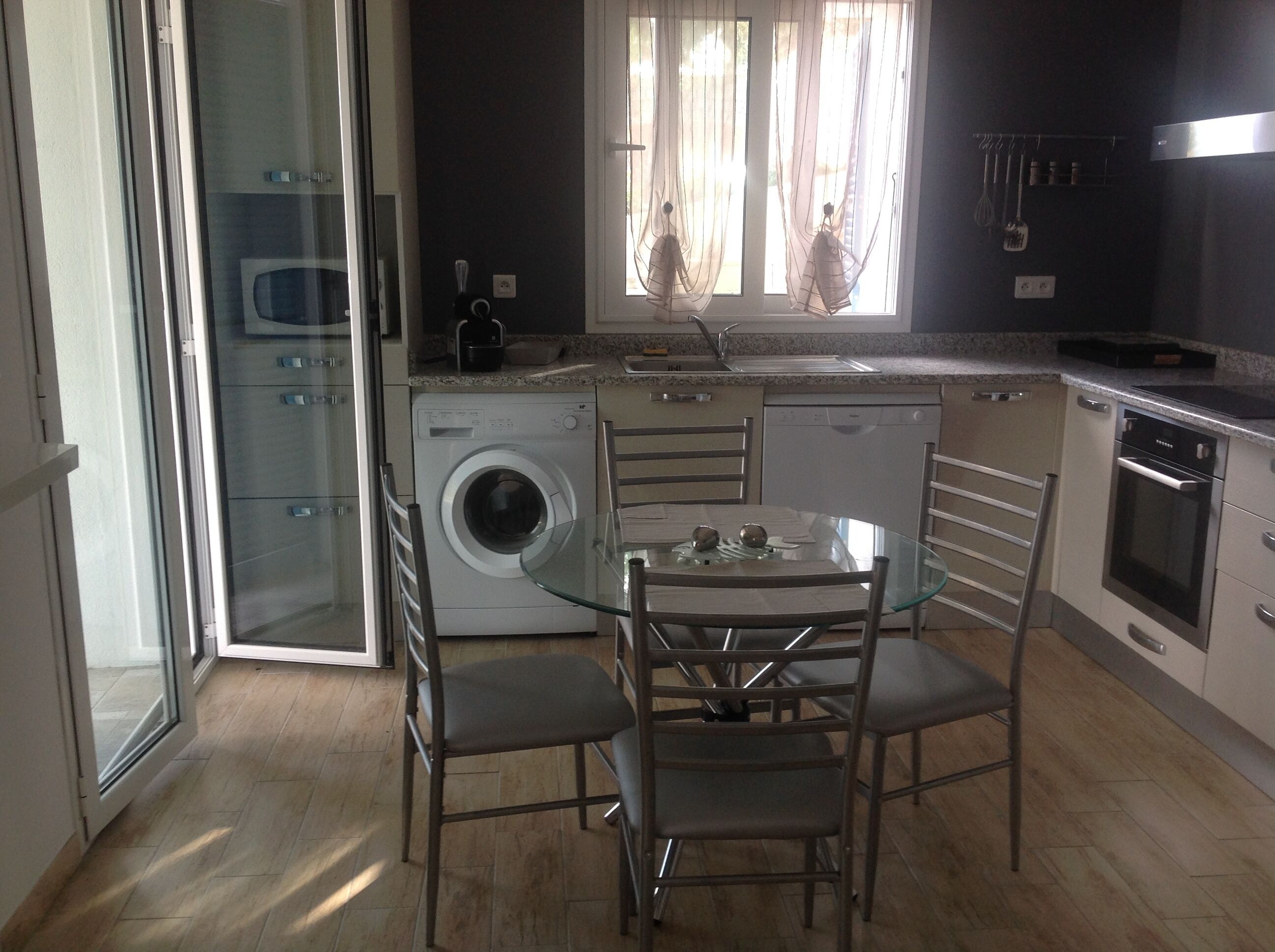 A compact kitchen with a washing machine, a dining table with chairs, and a window with sheer curtains.