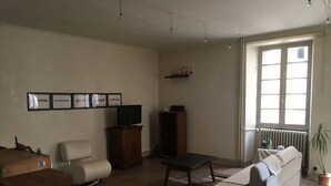 Una sala de estar con un sofá, una mesa de centro, un televisor sobre un mueble de madera, una silla, una mesita y un radiador cerca de una ventana.