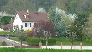A house with a sloped roof, a chimney, and a small balcony. There is a well-maintained lawn and a variety of trees, including some with white flowers.