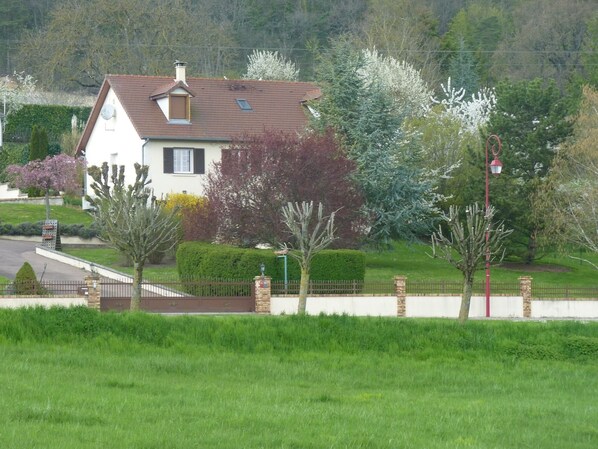 A house with a sloped roof, a chimney, and a small balcony. There is a well-maintained lawn and a variety of trees, including some with white flowers.