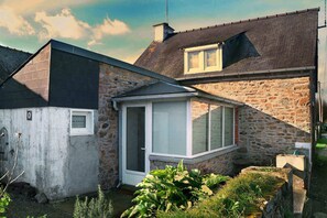Une maison en pierre avec un toit en pente, une petite dépendance attenante dotée d’une fenêtre, et un jardin planté.