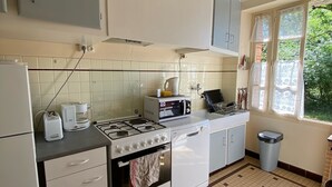 Une cuisine équipée d’une cuisinière à gaz, d’un four à micro-ondes et d’une machine à café. Une fenêtre ornée de rideaux en dentelle offre une vue sur la verdure environnante.