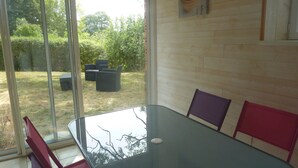 Une table à plateau en verre, quatre chaises, une vue sur un jardin par une porte-fenêtre coulissante et un mur en bois orné d’un tableau.