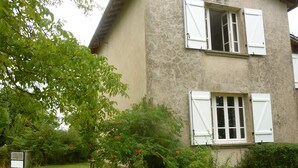 Une maison à deux étages avec des volets blancs, entourée d’une pelouse et de divers arbustes.