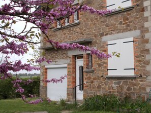 Un edificio in pietra con una porta bianca e persiane, circondato da un giardino con alberi in fiore rosa.