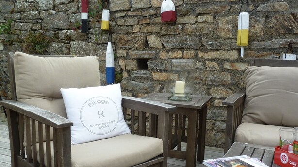 Une terrasse en bois avec deux fauteuils rembourrés, une table en verre et un mur de pierre orné de drapeaux colorés.