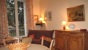 A cozy living room with a brown sofa, a wooden cabinet, a dining table with chairs, and a window with curtains.