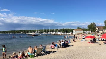 Une plage où des gens se détendent, certains sur le sable et d’autres dans l’eau. On aperçoit des bateaux et un quai au loin.