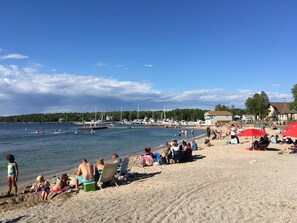 Una playa con gente relajándose, algunos en la arena y otros en el agua. Se ven barcos y un muelle al fondo.