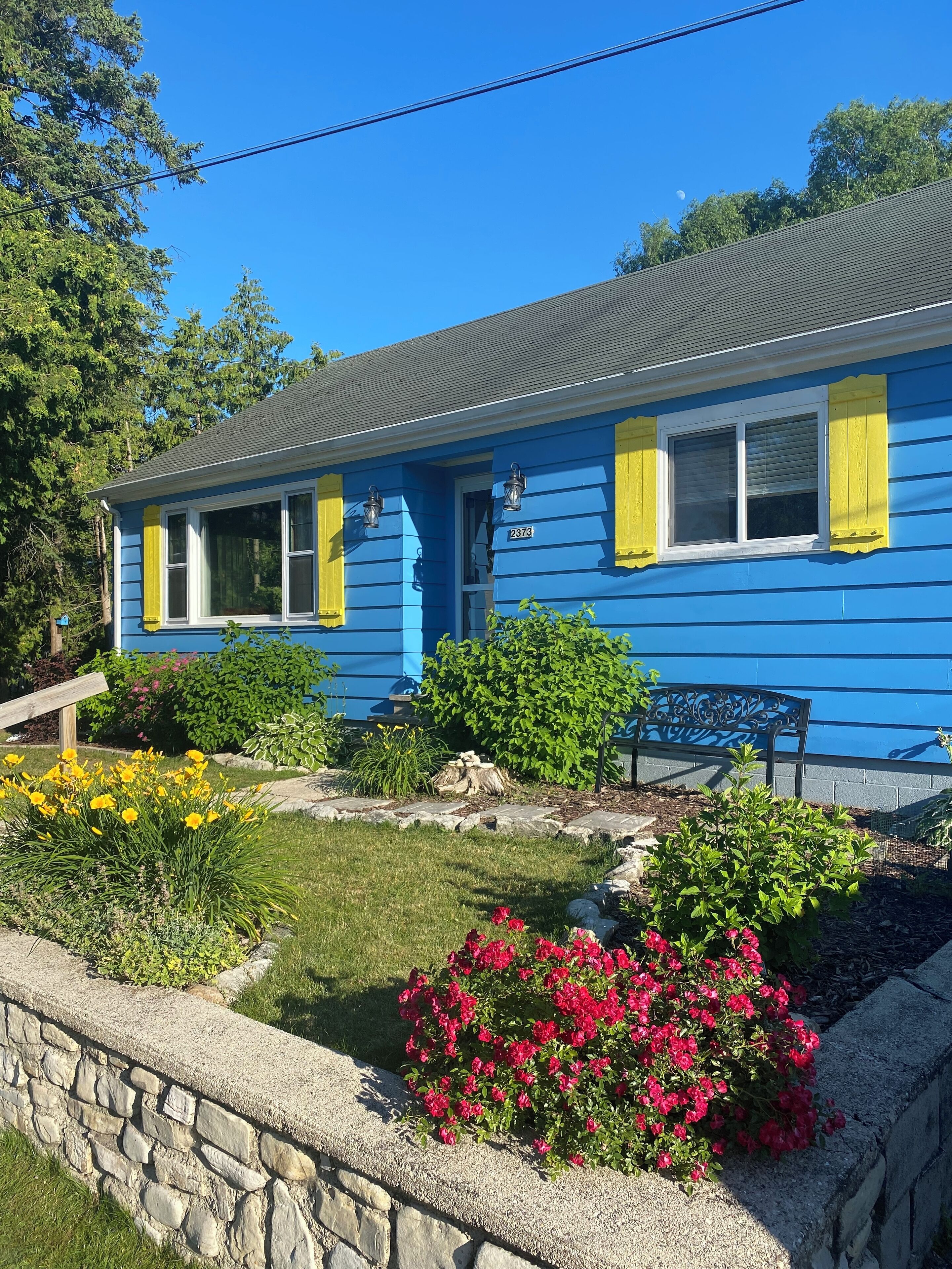 Une maison bleue avec des volets jaunes, un mur de pierre et un jardin fleuri.