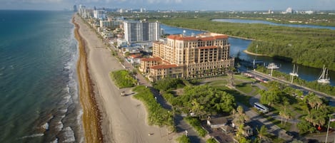 An aerial view of a coastal area with a beachfront hotel, a marina, and surrounding greenery.