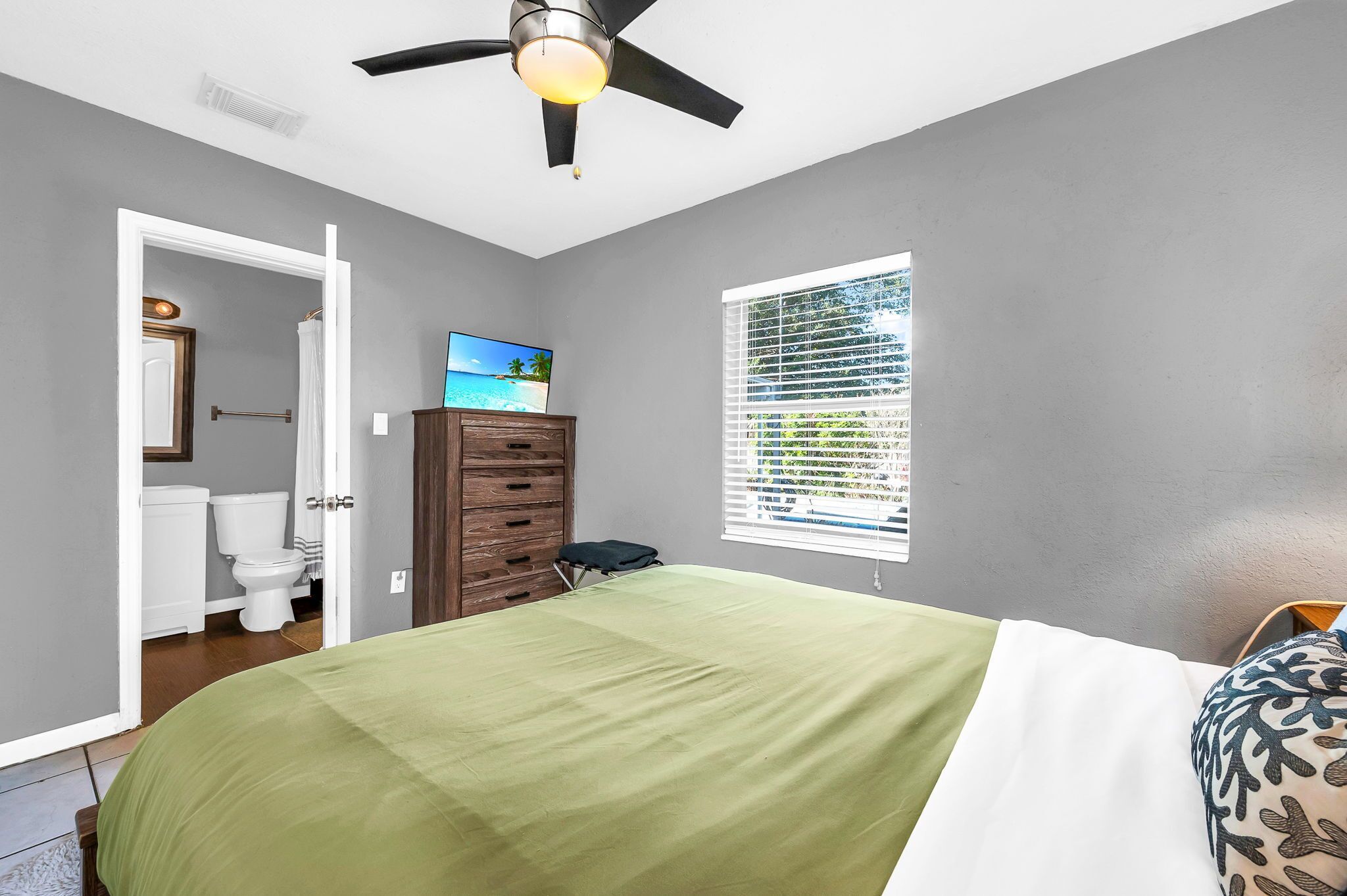 A bedroom with a bed, a ceiling fan, a window with blinds, a wooden dresser, and a bathroom visible through an open door.