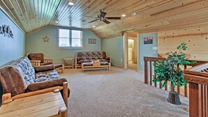 A cozy living room with a high ceiling, wooden ceiling beams, a ceiling fan, a large window, a sofa, a coffee table, and a staircase leading to an upper level.
