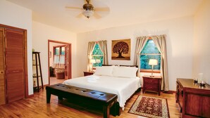 A bedroom with a large bed, wooden floor, a ceiling fan, a nightstand, a bedside table, a lamp, a window with curtains, and a wall-mounted artwork.