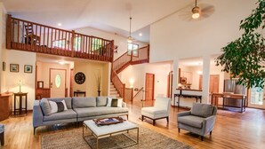 A spacious living room with a large gray sofa, a white armchair, and a coffee table. A wooden staircase leads to an upper level with a balcony.
