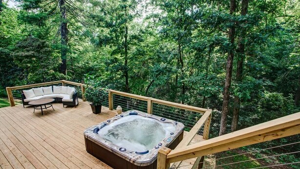 A wooden deck with a hot tub, a cushioned seating area, and a view of a forested area.