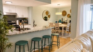 Une cuisine moderne avec un plan de travail en marbre, un coin repas avec une table et des chaises en bois, et un salon avec un canapé et des tabourets verts.