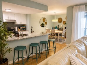 A modern kitchen with a marble countertop, a dining area with a wooden table and chairs, and a living area with a sofa and green stools.