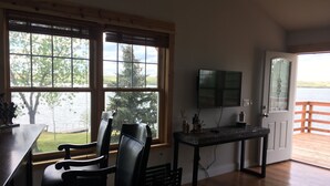 Una habitación con piso de madera, un escritorio con sillas, televisión y una puerta que da a un balcón con vista al lago.