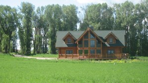 A log cabin with a large porch and multiple windows, surrounded by a grassy field and dense trees. - Yellowstone, Grand Teton, Fish, Ski, Parks, Pets, Bike, Fire Pit, (Freedom)