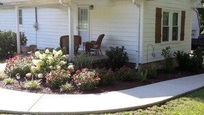 Una casa pequeña de color blanco con un porche, dos sillas y un jardín con flores rojas y blancas.