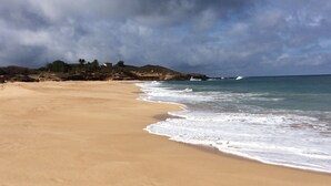 A sandy beach with waves lapping at the shore, a rocky outcrop in the background, and a cloudy sky. - Romantic Modern Upstairs Corner Studio with Ocean View, on the Island of Molokai (Maunaloa)