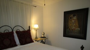 Een net opgemaakt bed met een witte spreidek en twee rode kussens, een bedlampje, een klein tafeltje met een boek en een buste, en een lijst met een schilderij aan de muur.