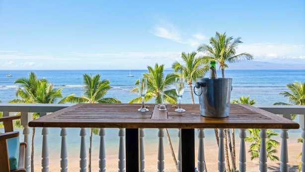 Een houten tafel met een metalen emmer en twee glazen op een balkon met uitzicht op een strand, palmbomen en de oceaan.