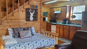 Interior de una cabaña con escalera de madera, cama con ropa de cama estampada, zona de cocina con armarios de madera y un refrigerador.