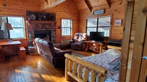 Interior acogedor de una cabaña con chimenea de piedra, muebles de madera, un televisor y ventanas amplias.
