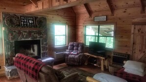 Interior acogedor de una cabaña con chimenea de piedra, un sillón cómodo, un televisor sobre una mesa de madera y un ventanal con vistas al exterior.