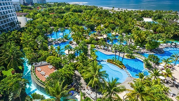 Un resort con múltiples piscinas rodeado de exuberante vegetación y palmeras, con vista a una playa y el océano.
