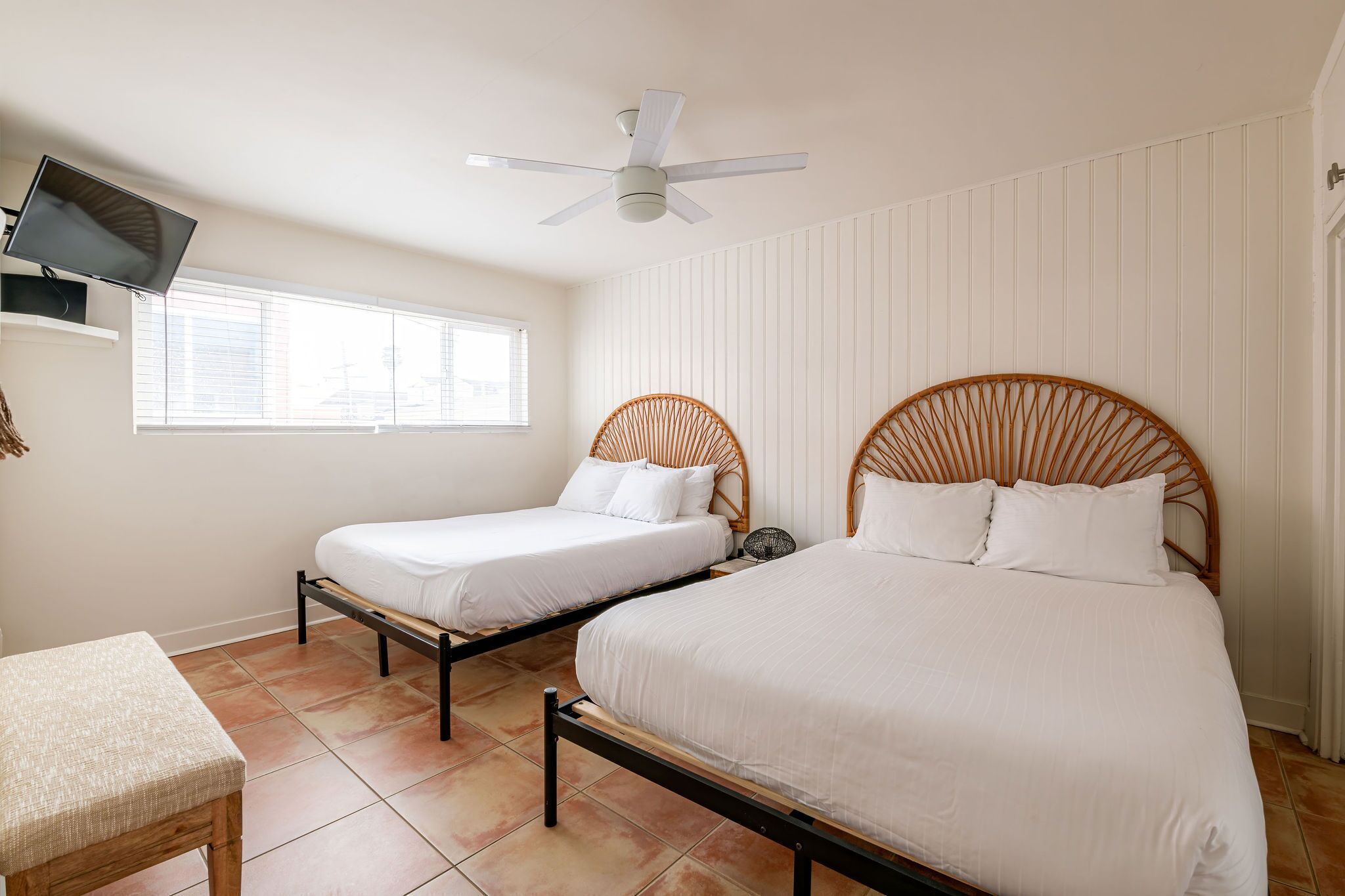 Une chambre avec deux lits simples, un ventilateur de plafond, un téléviseur fixé au mur et une fenêtre avec des stores.