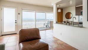 Un salon moderne avec un canapé en cuir, un coin cuisine agrémenté d’un miroir rond, et un balcon offrant une vue sur la mer.