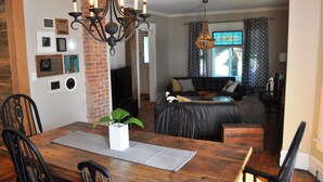 A dining area with a wooden table, black chairs, and a chandelier. There is a brick wall with framed pictures and a television in the background.