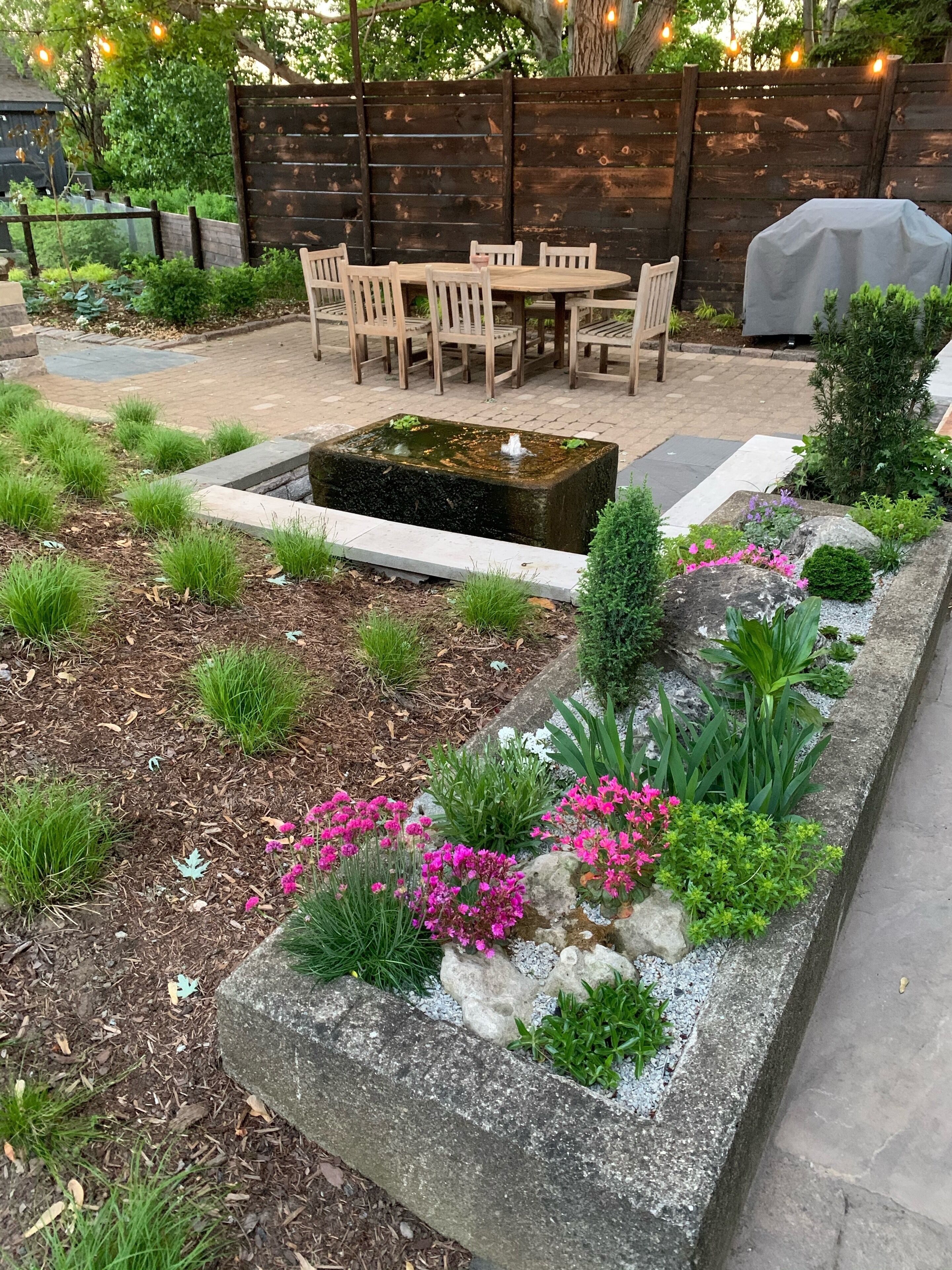 Un jardín con una fuente, sillas de madera y una zona de barbacoa.