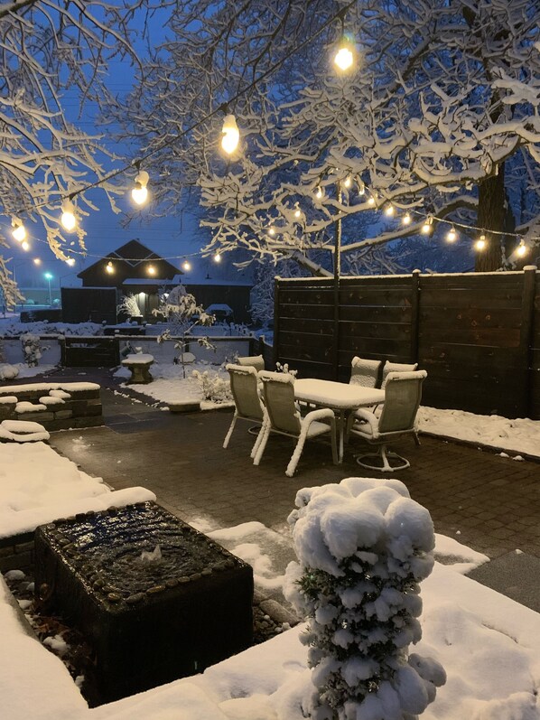 An outdoor patio area with a table and chairs, surrounded by snow-covered plants and a wooden fence.