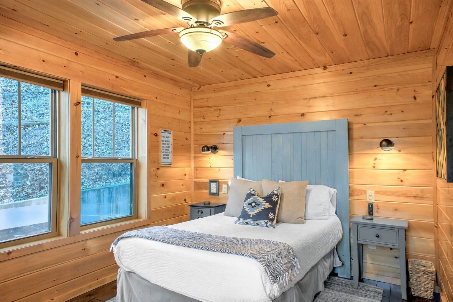 A wooden cabin bedroom with a bed, nightstands, a ceiling fan, and a window.