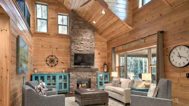 A log cabin interior with a stone fireplace, wooden walls, and a large window overlooking trees. - FishTop Cabin | Luxury Mountain Cabin Across from the Green River (Saluda)