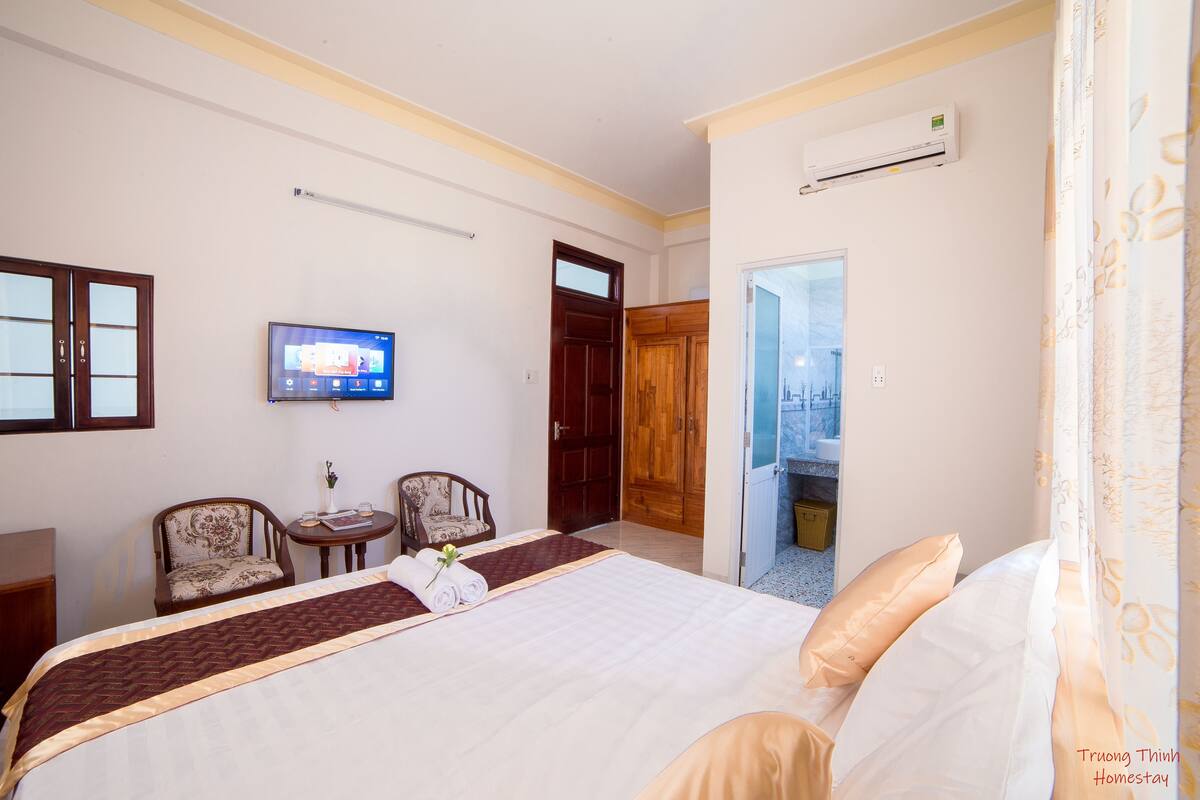 A hotel room with a bed, two chairs, a TV, and an air conditioning unit.