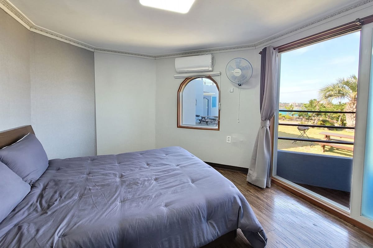 Ein Schlafzimmer mit Bett, Fenster, Ventilator und Blick ins Freie.