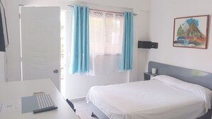 Habitación de hotel con cama, escritorio, silla, ventana con cortinas, lámpara de pared y un cuadro.