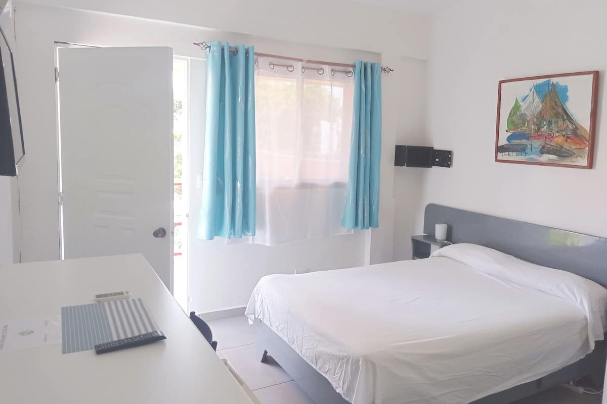 Une chambre d’hôtel comprenant un lit, un bureau, une chaise, une fenêtre avec des rideaux, une lampe fixée au mur et un tableau.